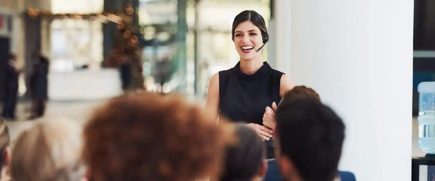 A young woman speaking in front of an audience using a headset and attached microphone.
