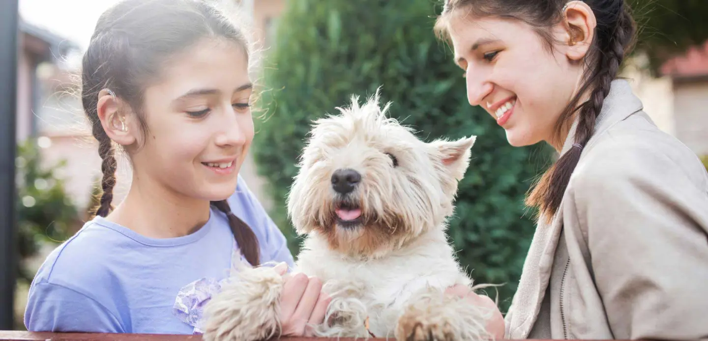 Two girls with hearing aids playing with their dog.