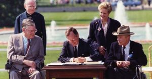 President George HW Bush signs the ADA bill on the White House lawn with two men in wheelchairs on either side of the table