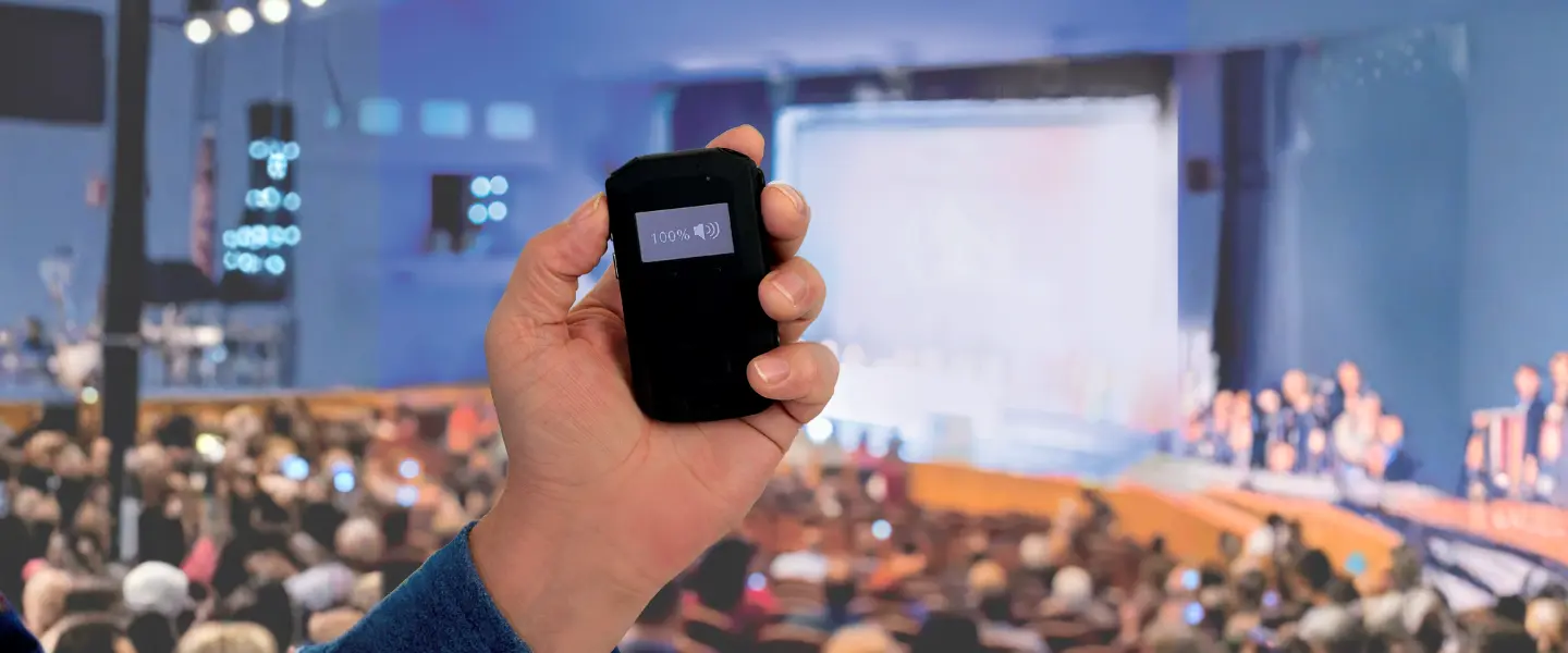 A close up of a hand holding an assistive listening device with Auracast. A crowd is blurred out in the background.