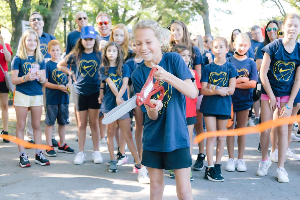 A young girl, Nikki Kramer, cuts the ribbon of a Walk4Hearing event with giant scissors, in front of a team of family and friends
