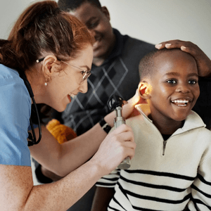Female audiologist uses an otoscope to examine the ear of a smiling young boy while his father watches.
