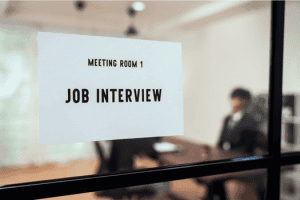 Sign on a conference room window reads "Meeting Room 1 - Job Interview" with a blurred job candidate in the background