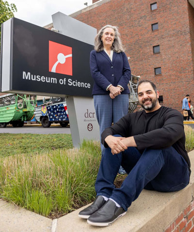 An older woman stands above a younger man seated on a brick wall outdoors, in front of a Museum of Science sign.
