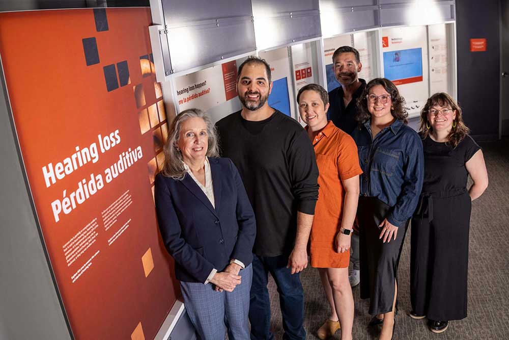 Group of six diverse people in front of a bilingual sign that reads "Hearing loss" in English and "Perdida auditiva" in Spanish.