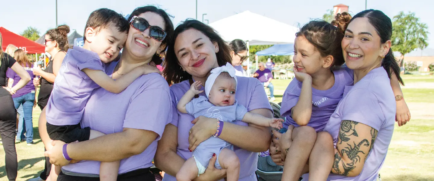 Group of women and children posing together at an outdoor event, all wearing matching lavender shirts and smiling at the camera.
